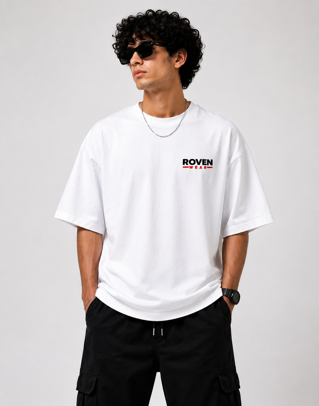 A man wearing a white oversized short-sleeve t-shirt with a small black and red 'ROVEN' logo on the chest, paired with black cargo pants, sunglasses, and a silver chain necklace, photographed against a light gray studio background.