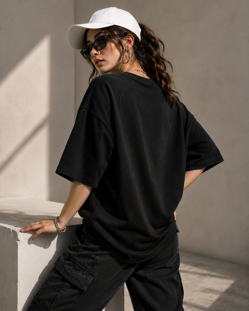 A woman wearing an oversized black t-shirt, black cargo pants, a white baseball cap, and sunglasses, posing outdoors against a textured concrete wall.